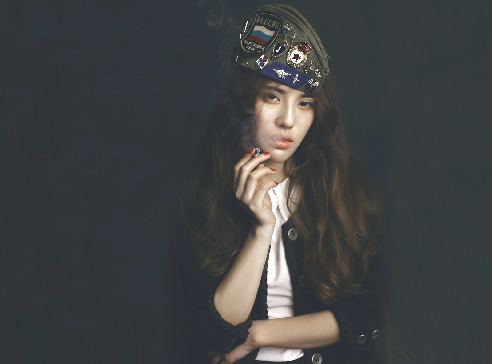 긴 머리의 여성이 장식 배지가 달린 베레모를 쓰고 어두운 배경 앞에서 손을 얼굴 가까이에 댄 채 포즈를 취하고 있다.