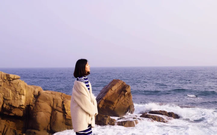 한 여성이 바위 해안에 서서 바다와 부서지는 흰 파도를 바라보고 있다.