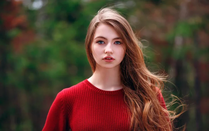 一名长发女子穿着红色针织衫站在绿色森林背景前。