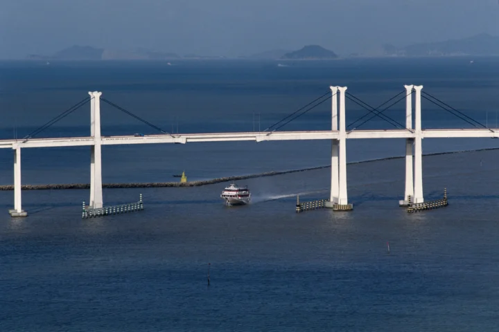 一座白色桥梁横跨蓝色海面，桥下有一艘小船驶过。