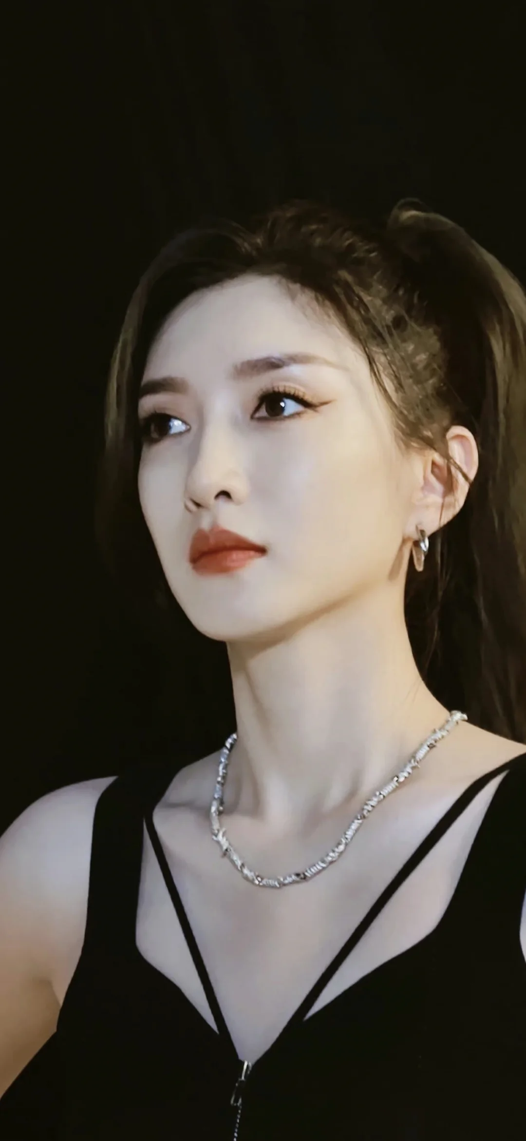 黒いドレスを着た成人女性が、暗い背景の前で横顔を見せながら横を見つめている。