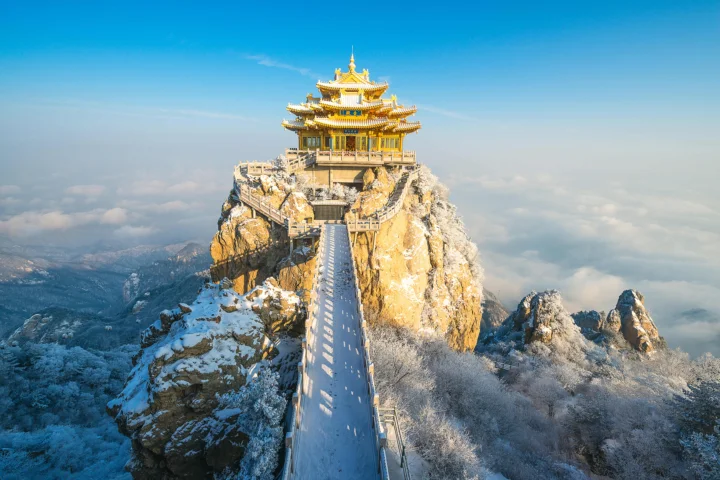 一座金色传统楼阁坐落在被积雪覆盖的山峰之上，远处云海与湛蓝天空相映成景。