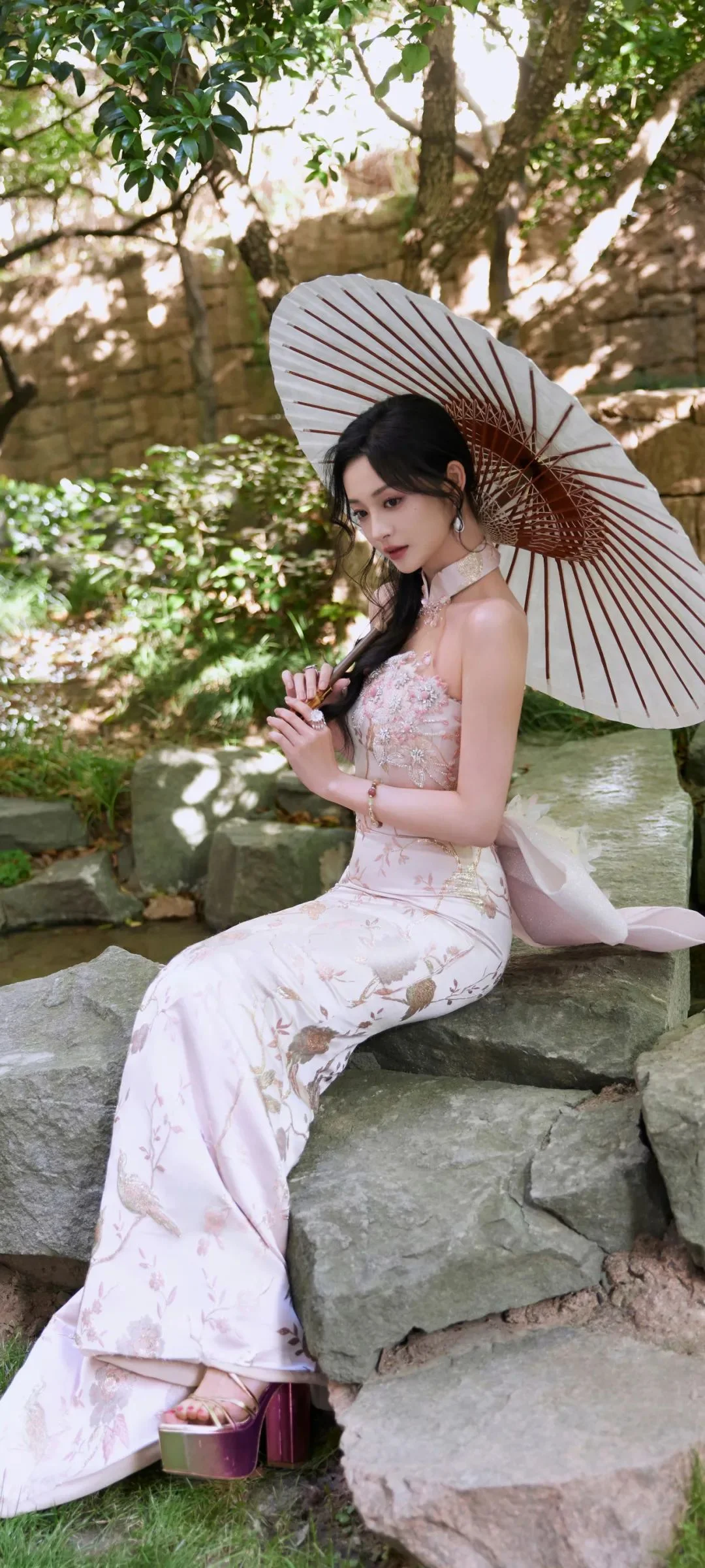 一名身穿淺色傳統服飾的成年女性坐在庭院石階上，手持油紙傘，周圍是樹木與岩石。