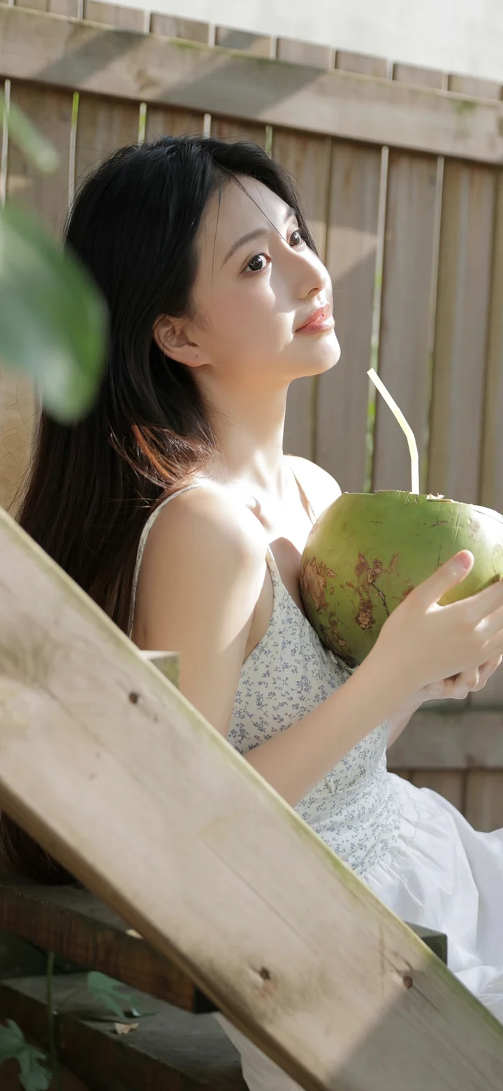 一名成年女性坐在木制栏杆旁，手捧一只青椰子沐浴在阳光中。
