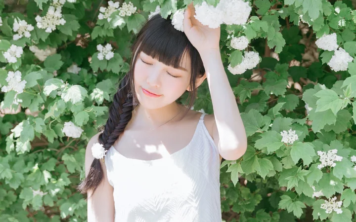 一名女子闭着眼站在绿叶和白色花朵环绕的户外环境中。