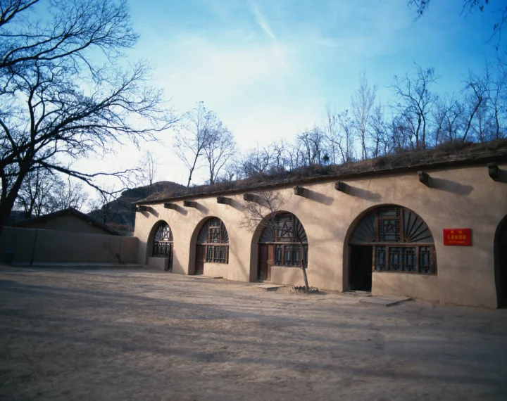 一座黄土色古建筑立于铺装庭院旁，墙面设有拱形窗户，周围可见冬季的无叶树木和蓝天。