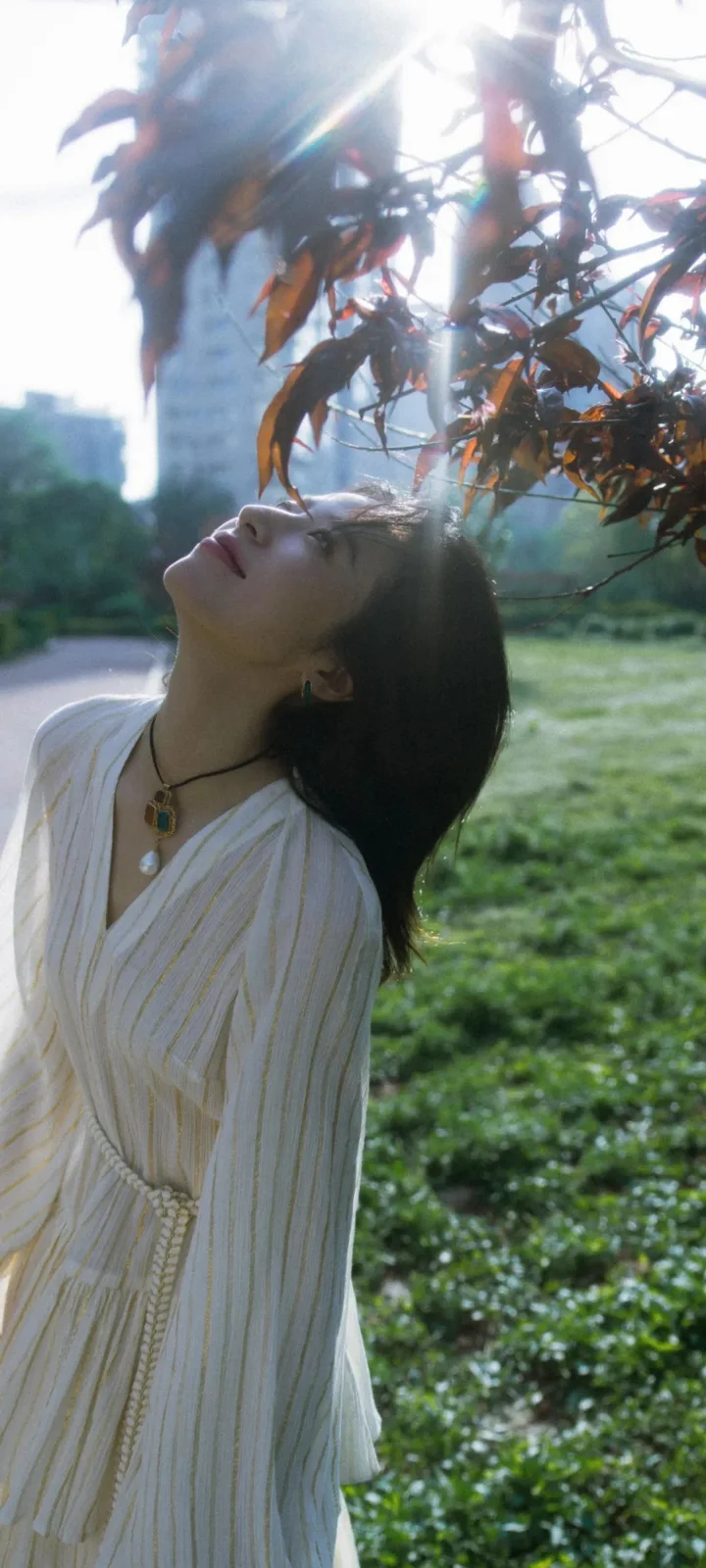 一名身穿白色长裙的女性在树枝和阳光下抬头站立，背景是草地与模糊的建筑。