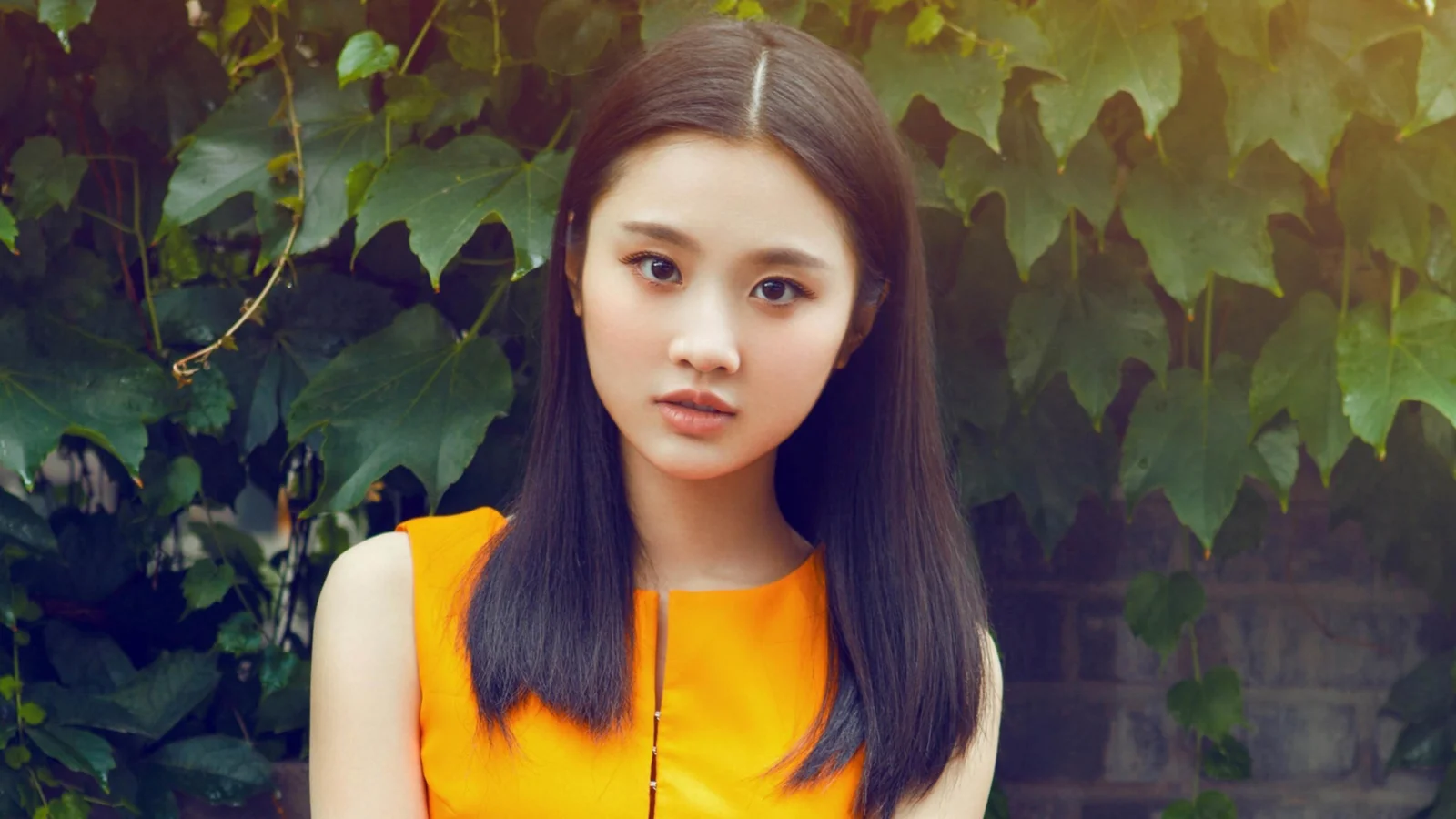 一名长发女性穿着橙色上衣站在绿叶背景前直视镜头。