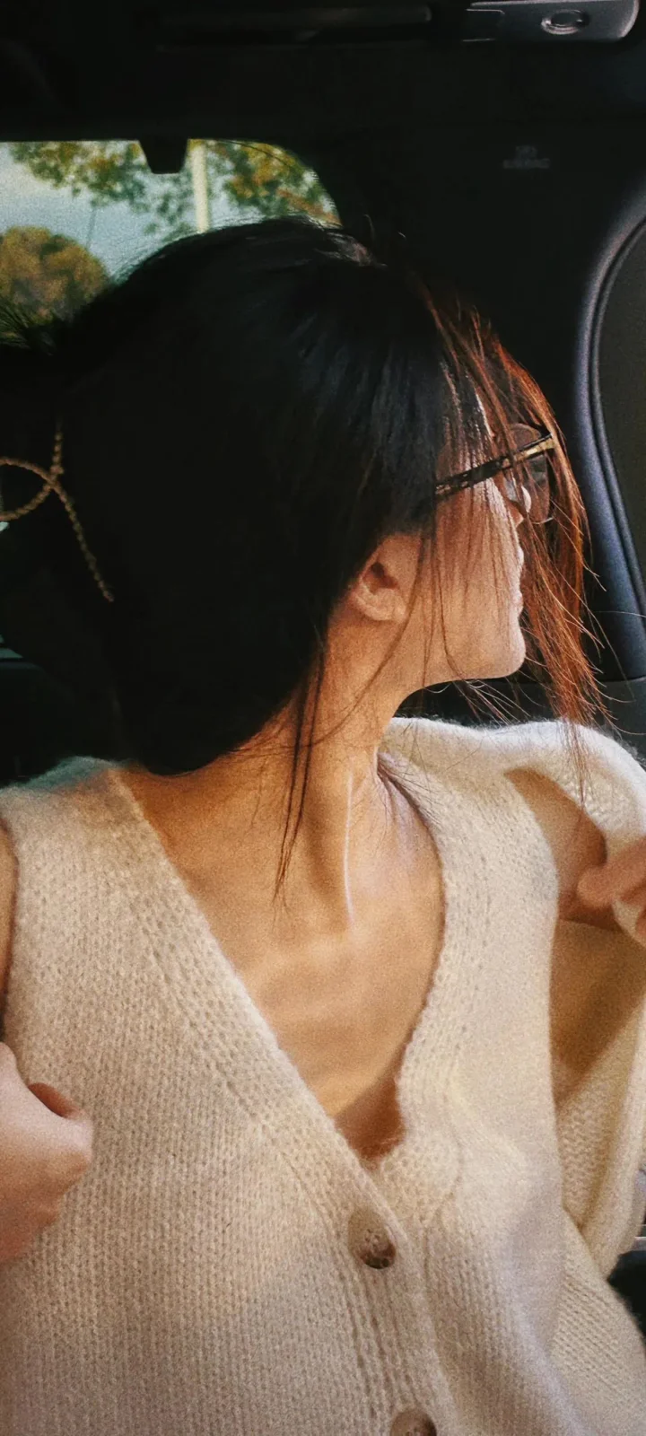 一名成年女性坐在车内，穿着米白色针织开衫并侧头望向车窗外。