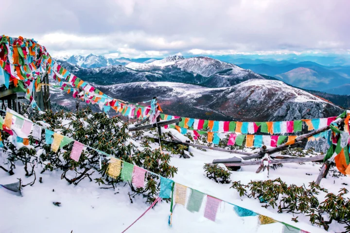 彩色经幡悬挂在积雪山坡前，远处是连绵的雪山和阴云天空。