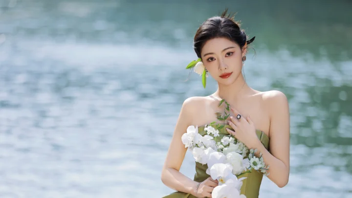 一名女子站在水边，手捧一束白色花朵，背景是柔和的水面。