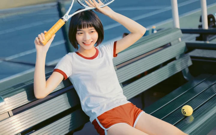 短い髪の女性がベンチに座り、テニスラケットを持ち、隣にテニスボールが置かれています。
