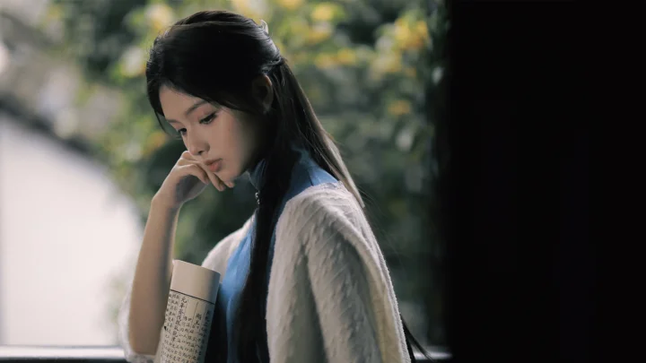 긴 머리의 여성이 창가에서 책을 들고 옆을 바라보고 있다.