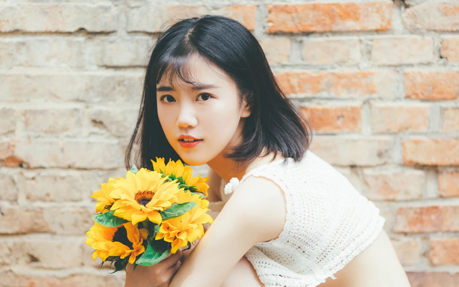 黒髪の女性がレンガの壁の前でひまわりの花束を抱えている。