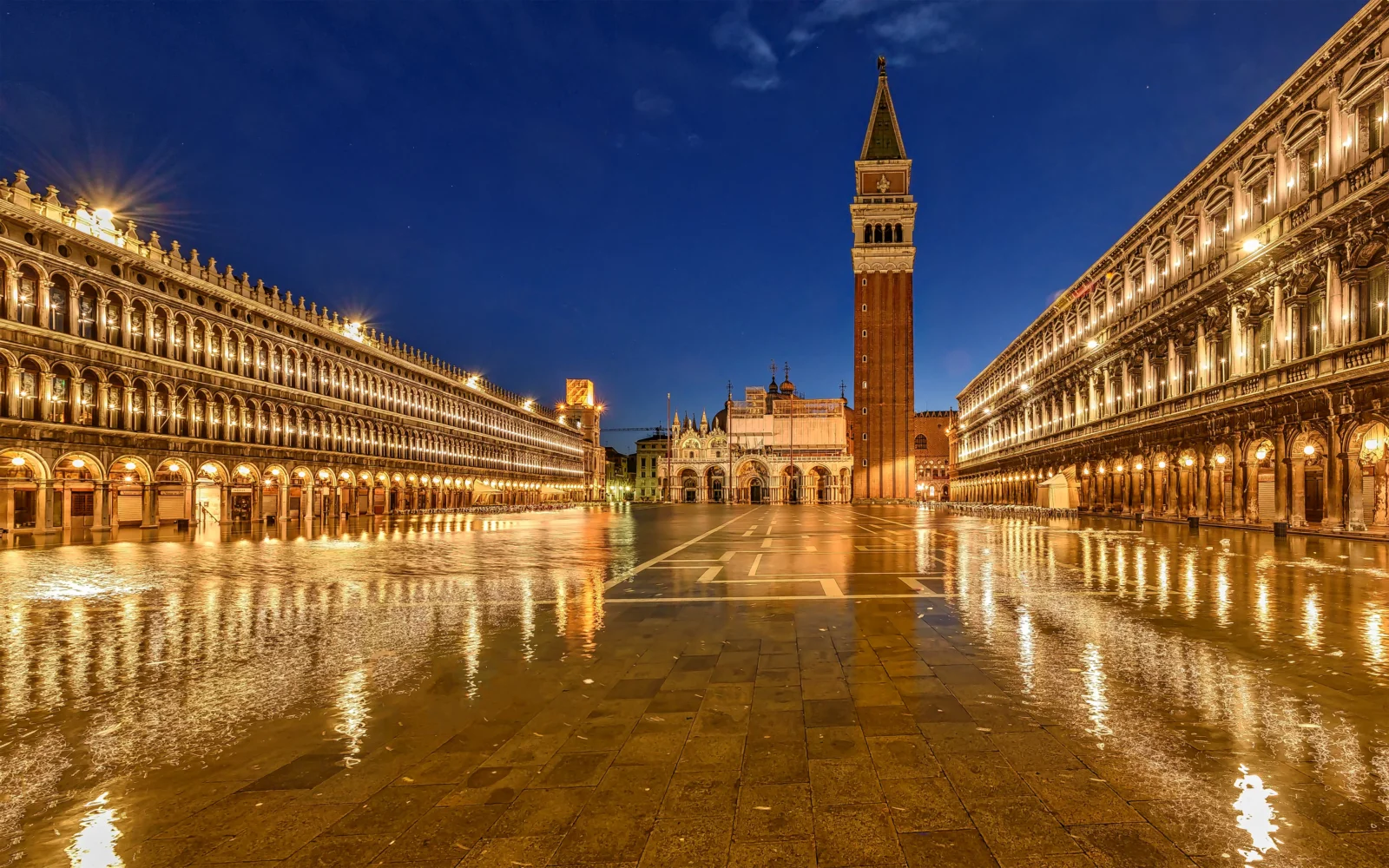 St. Mark's Square in Venice at dusk, wet pavement reflecting the colonnades and campanile.