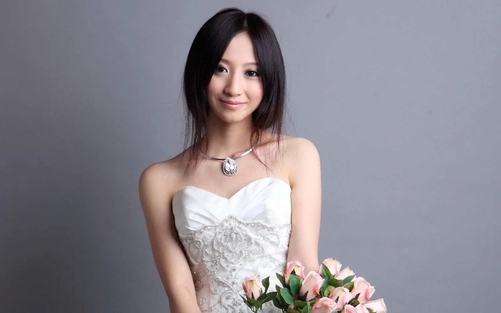 白いドレスを着た笑顔の成人女性が、灰色の背景の前でピンクの花束を持っています。