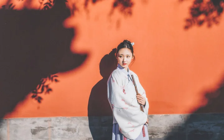 一名身穿浅色传统服饰的女子站在橙色墙前，墙面映出树叶状阴影。