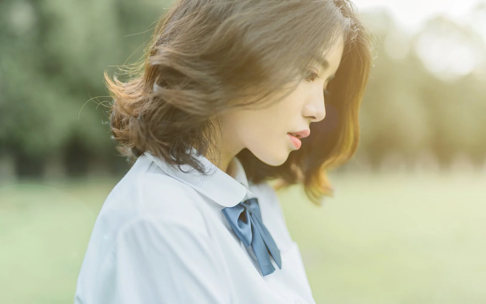 단발머리 여성이 야외의 부드러운 햇살 속에서 옆모습을 보이며 서 있다.