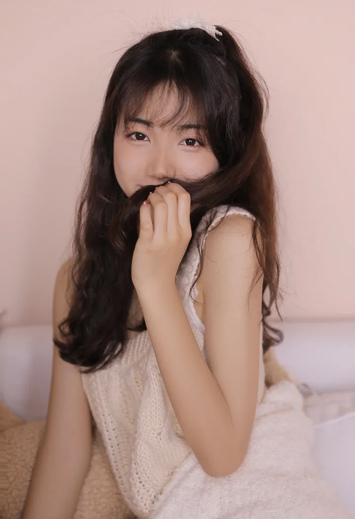 부드러운 분홍색 배경 앞에서 긴 머리의 여성이 옆모습으로 카메라를 바라보고 있다.
