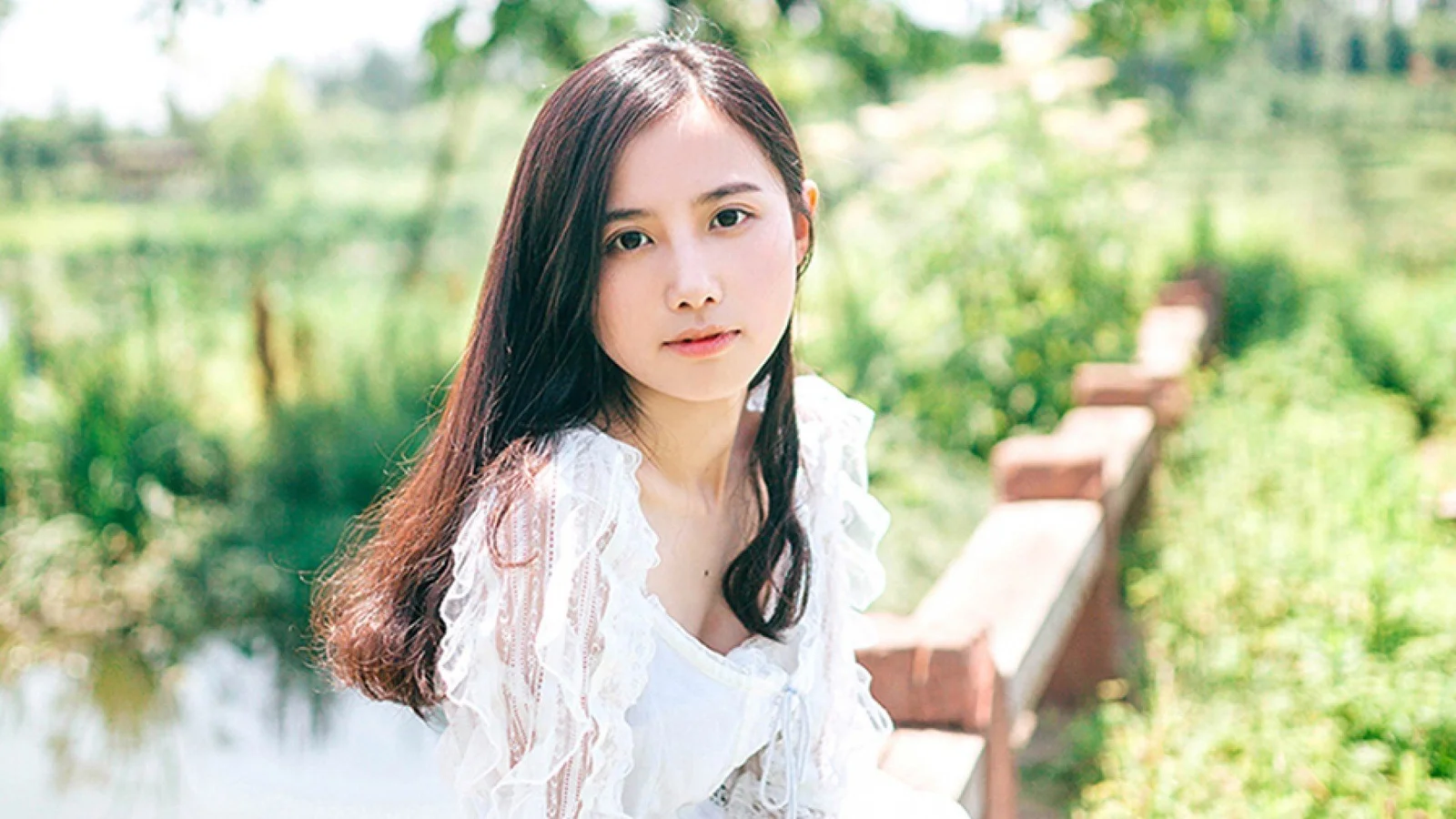 一名长发女子穿着白色上衣站在绿意盎然的水畔木桥旁。