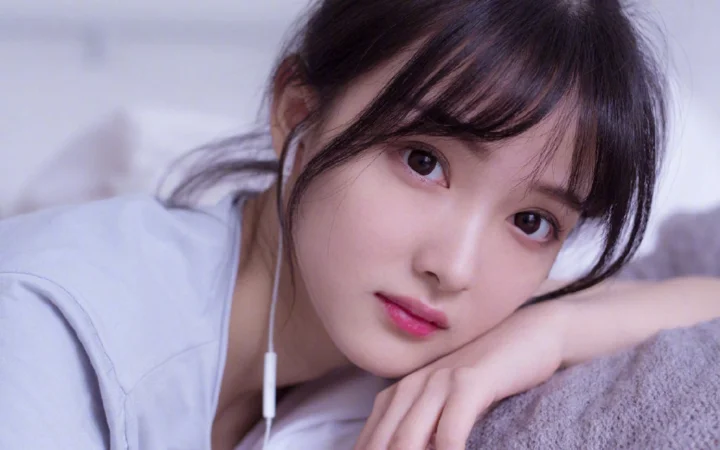 一名女子穿着白色上衣，戴着有线耳机，靠在柔软表面上看向镜头。