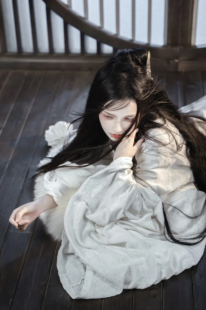 一名身穿白衣、留着长发的女子坐在木地板上，窗边光线映照着她的侧脸。