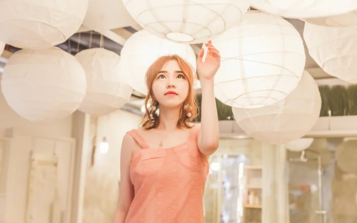 一名女性站在室内悬挂的白色纸灯笼下，抬手触碰上方的灯笼。