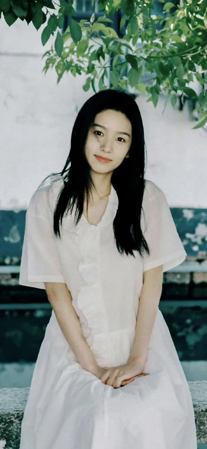 흰색 원피스를 입은 긴 머리 여성이 나무 아래에 앉아 카메라를 향해 미소 짓고 있다.