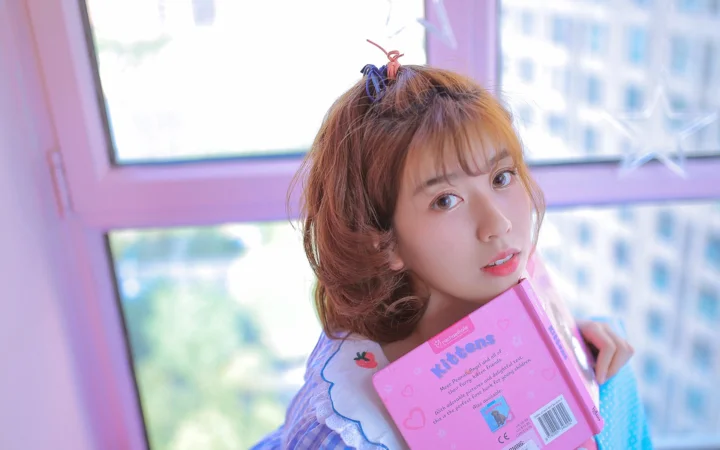 창가에서 분홍색 책을 든 젊은 여성이 카메라를 바라보고 있습니다.