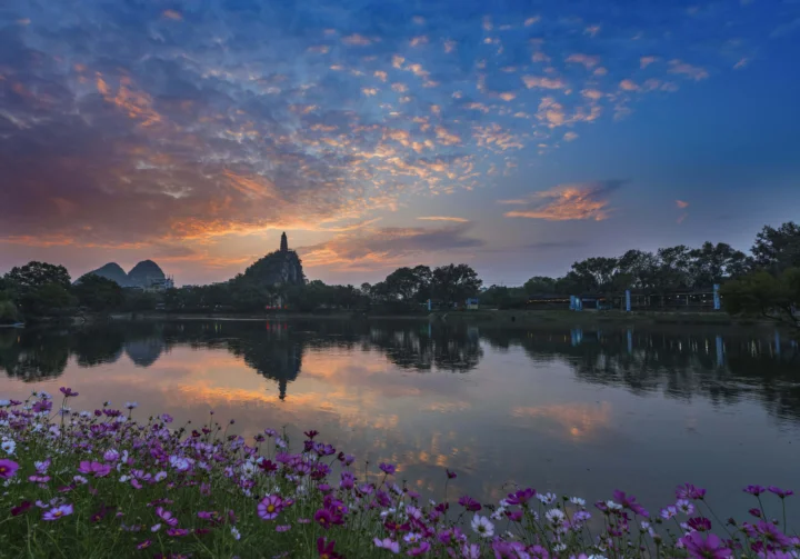 夕阳映照湖面，远处山峦与尖塔倒映在水中，岸边盛开着紫色花朵。
