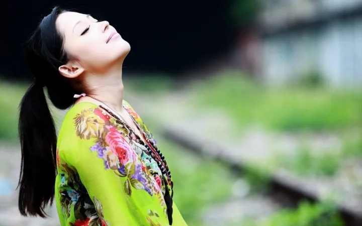 一名扎着马尾的女子身穿鲜艳花卉上衣，在户外仰头闭目，背景为虚化的绿色景物。