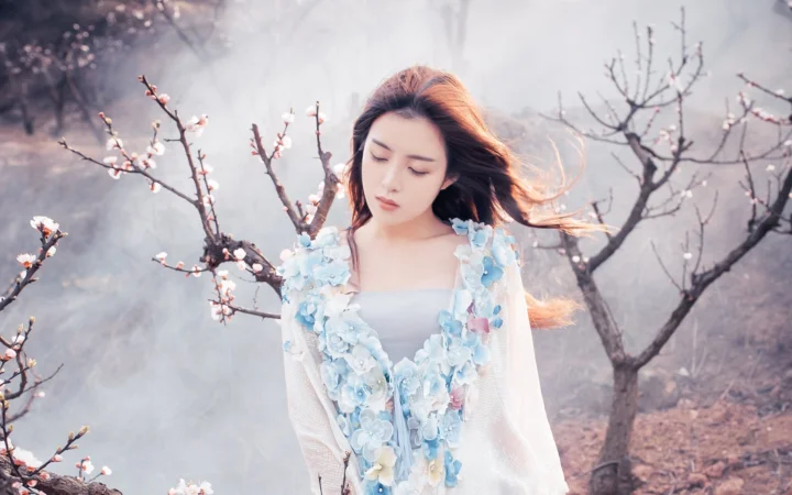 연한 파란색 꽃 장식 의상을 입은 여성이 부드러운 안개에 둘러싸인 꽃핀 나뭇가지 사이에 서 있습니다.