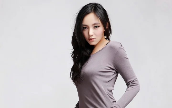 긴 머리의 여성이 회보라색 긴소매 상의를 입고 연한 회색 배경 앞에 서 있습니다.