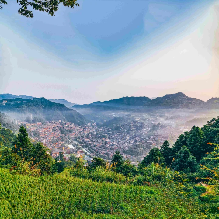 层叠山峦、森林和云雾环绕着山谷中的村寨，前景是一片绿色草地。