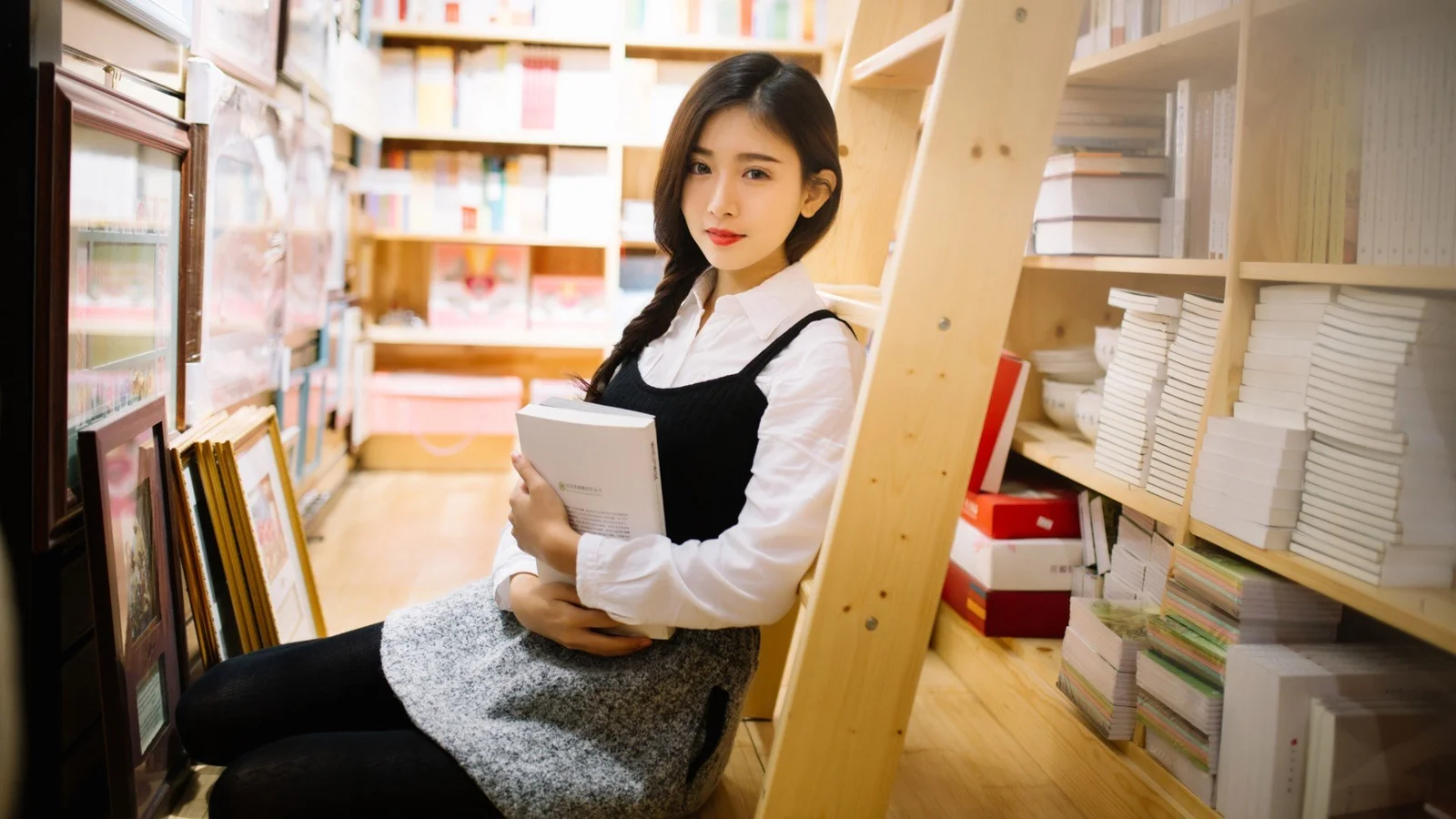 성인 여성이 책을 안고 서점 책장 사이에 앉아 있습니다.