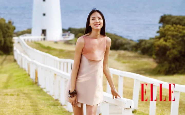 一名身穿浅粉色连衣裙的成年女子站在白色栅栏旁，背景是灯塔、草地和树木。