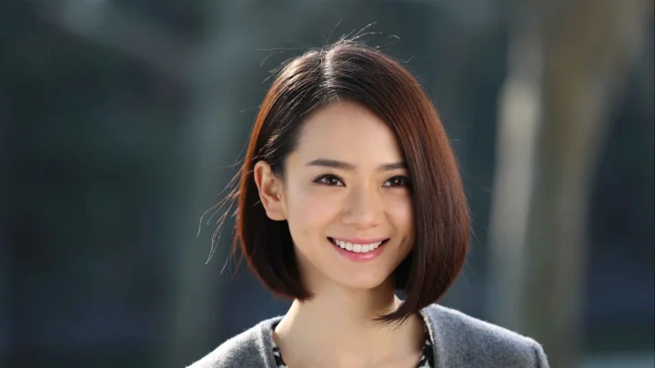 一位短棕发女性面带微笑，身穿灰色上衣，背景呈现柔和的深色虚化效果。