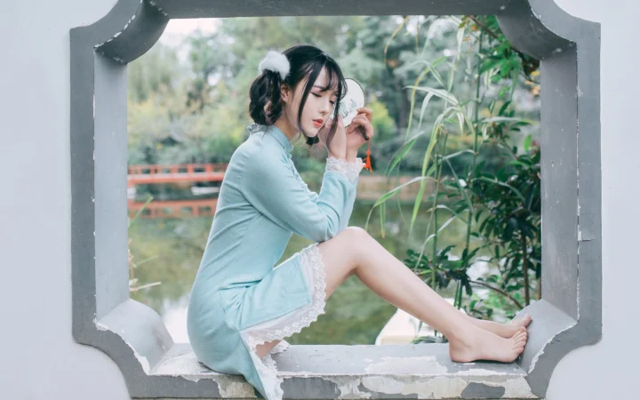 一名女子坐在花园景色前的窗台上，手持圆镜整理头发。