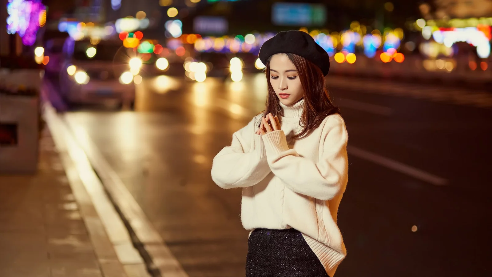 一名戴黑色贝雷帽、穿白色毛衣的成年女性站在夜晚灯光璀璨的城市街道旁。