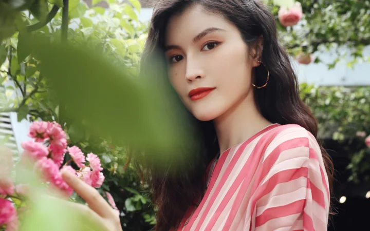 一名长发女性身穿粉色条纹上衣，站在盛开粉色花朵的花园中。