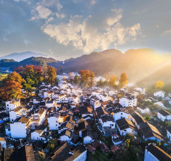 阳光洒在山间村落和层叠山岭上，白墙黑瓦建筑延伸至画面前景。