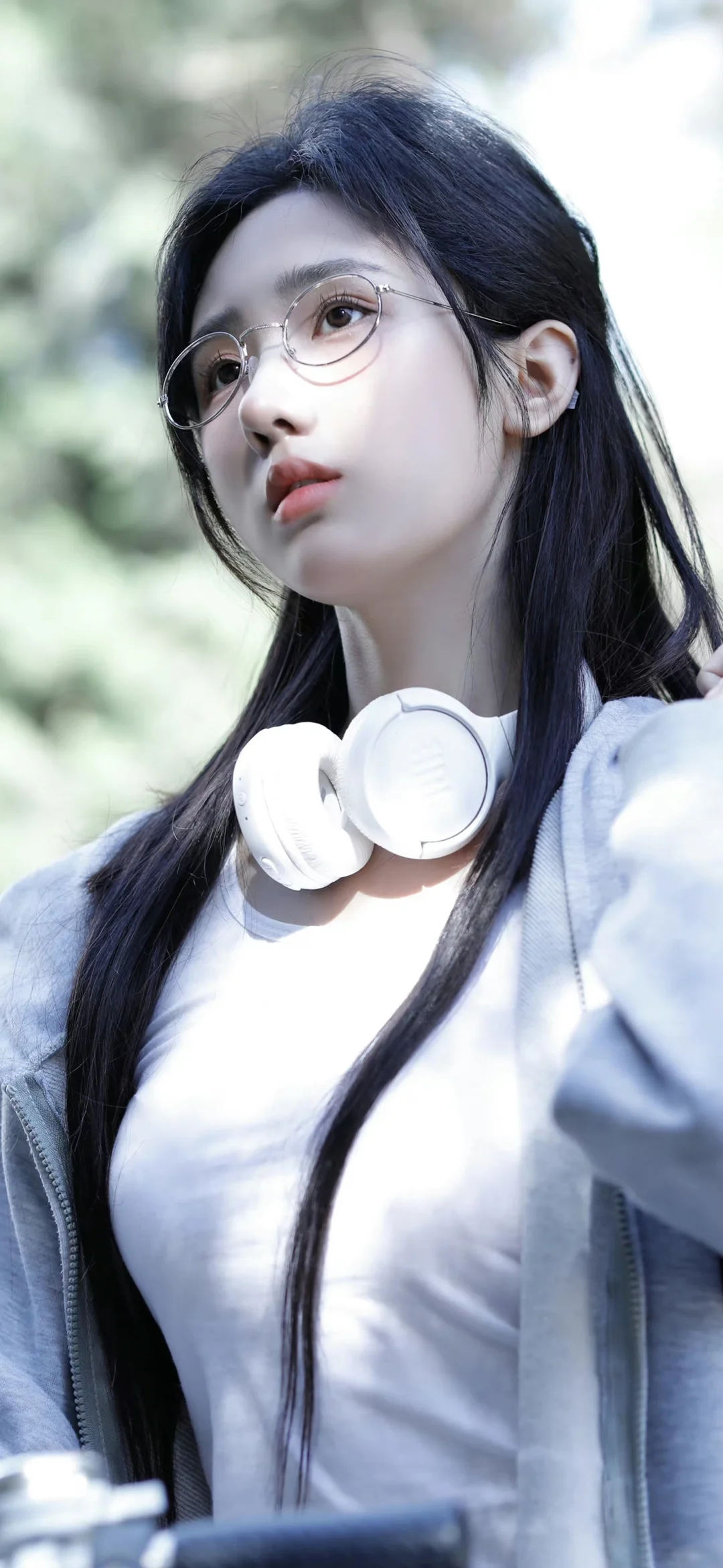 一名戴圆框眼镜的长发女性站在户外，白色耳机挂在颈部，背景为虚化的绿色植物。