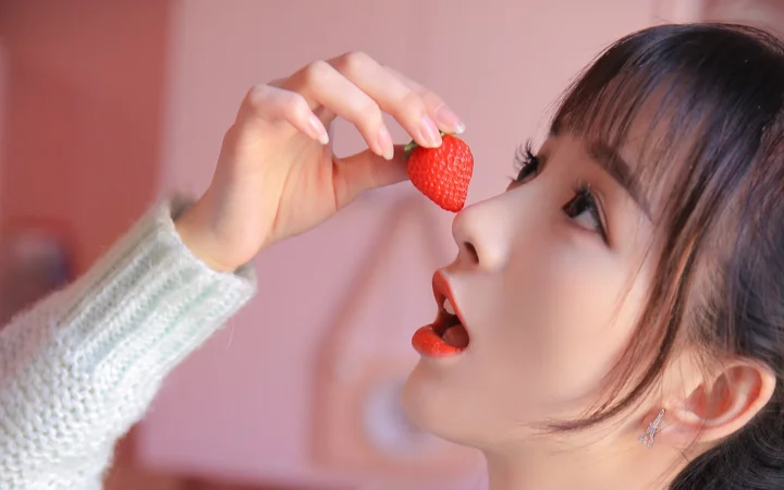 분홍색 배경 앞에서 여성이 딸기를 벌어진 입 가까이에 들고 있다.