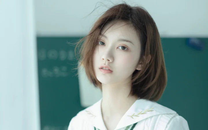 一位短发女性在柔和光线下直视镜头，背景为室内黑板。