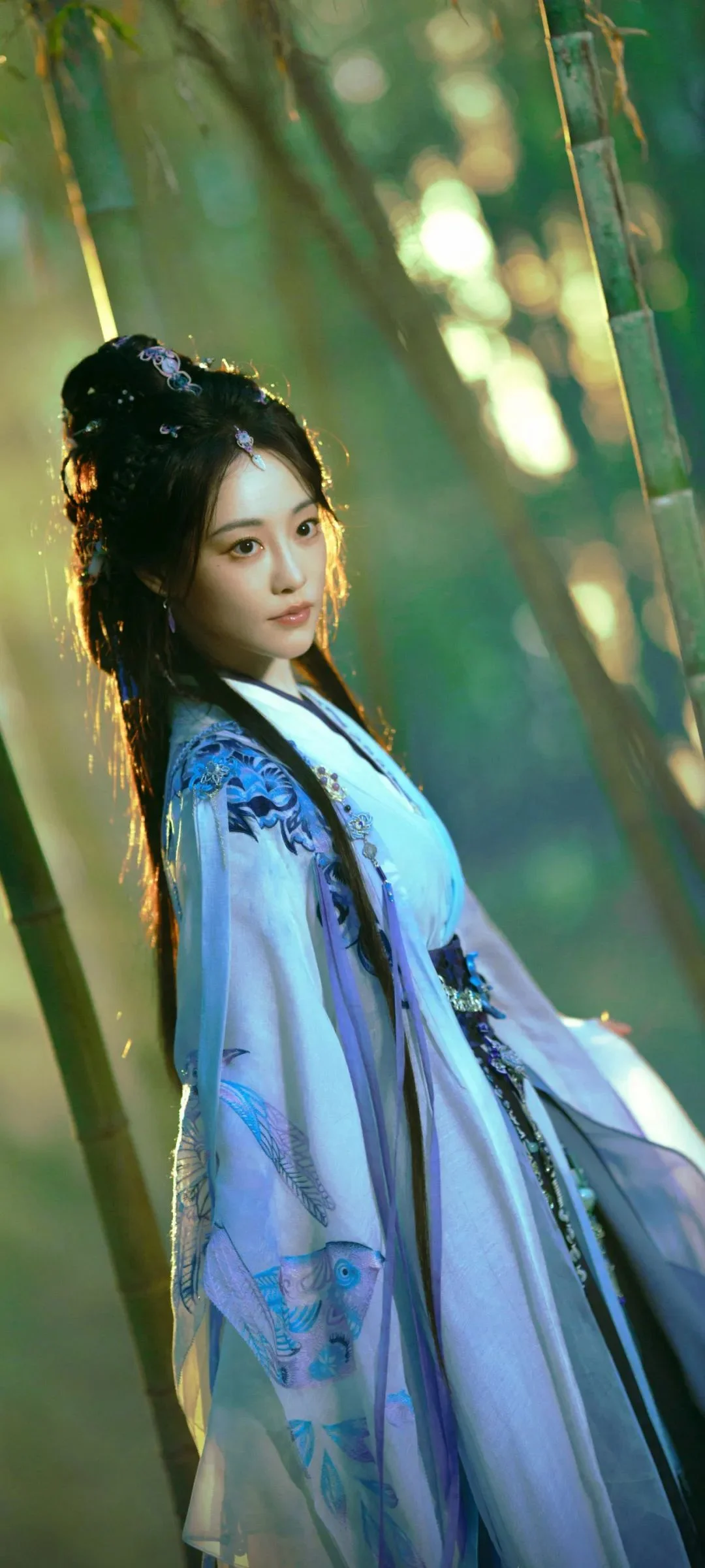 푸른 전통 의상을 입은 여성이 햇살이 비치는 숲에 서 있다.