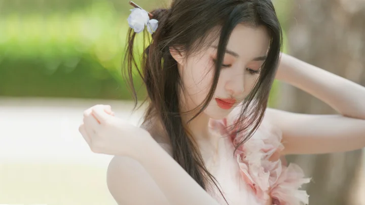한 여성이 부드럽게 흐릿한 야외 녹색 배경 앞에서 고개를 숙이고 머리에 흰 꽃을 꽂고 있다.