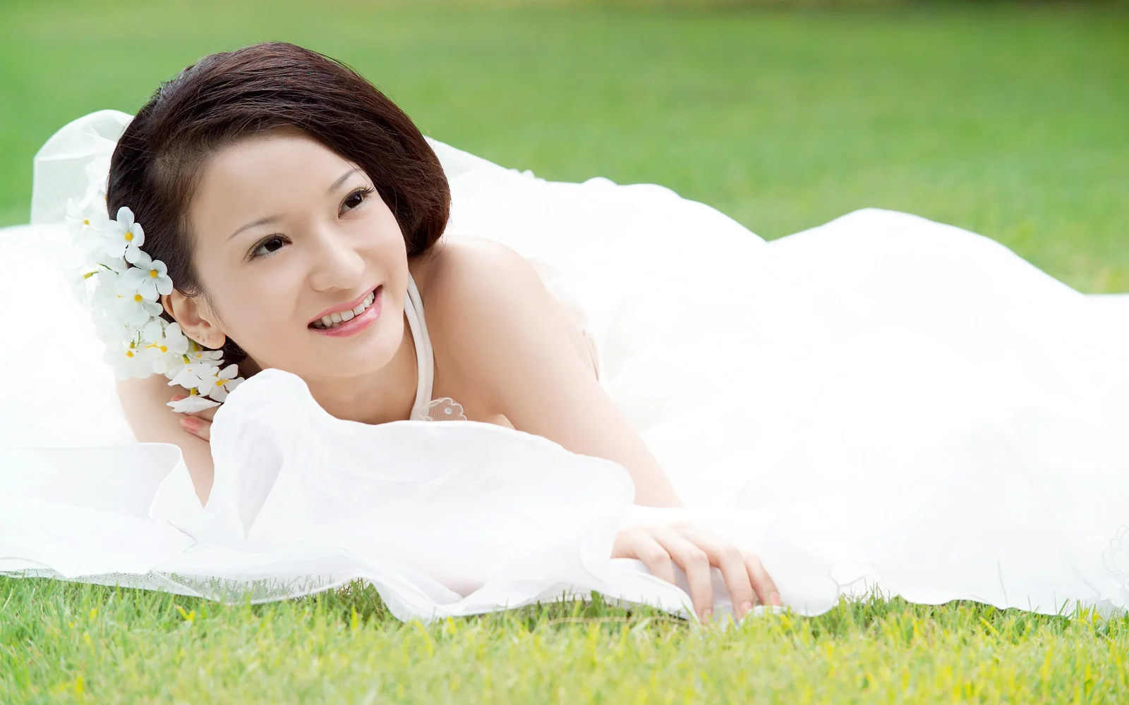 一名女子躺在草地上的白色床单上微笑，头发旁点缀着白色花朵。