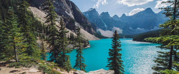 碧蓝湖泊 surrounded by evergreen forests and rugged mountains under a bright sky.