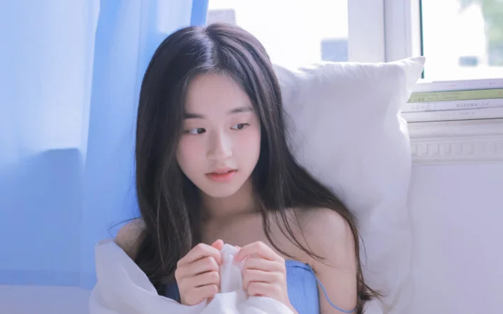 一名长发女性坐在窗边的床上，手持白色布料，画面以柔和的蓝白色调为主。