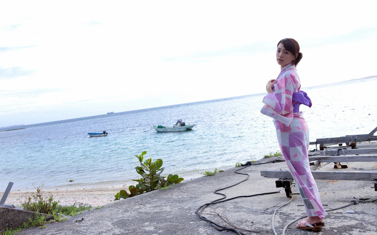 一名女子站在海边的水泥岸上，远处可见海面和小船。